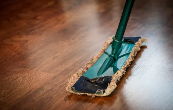 A close-up shot of a floor mop head with a green handle cleaning a glossy brown hardwood floor, symbolizing maintenance, property upkeep, and cleaning labor costs.