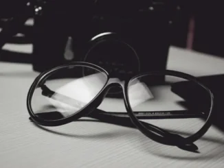 A close-up, black and white photo of a pair of thick-rimmed black glasses resting on a wooden surface in front of a vintage camera.