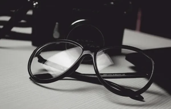 A close-up, black and white photo of a pair of thick-rimmed black glasses resting on a wooden surface in front of a vintage camera.