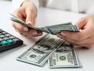 Close-up of a person's hands counting a stack of one hundred dollar bills on a white desk next to a black calculator.