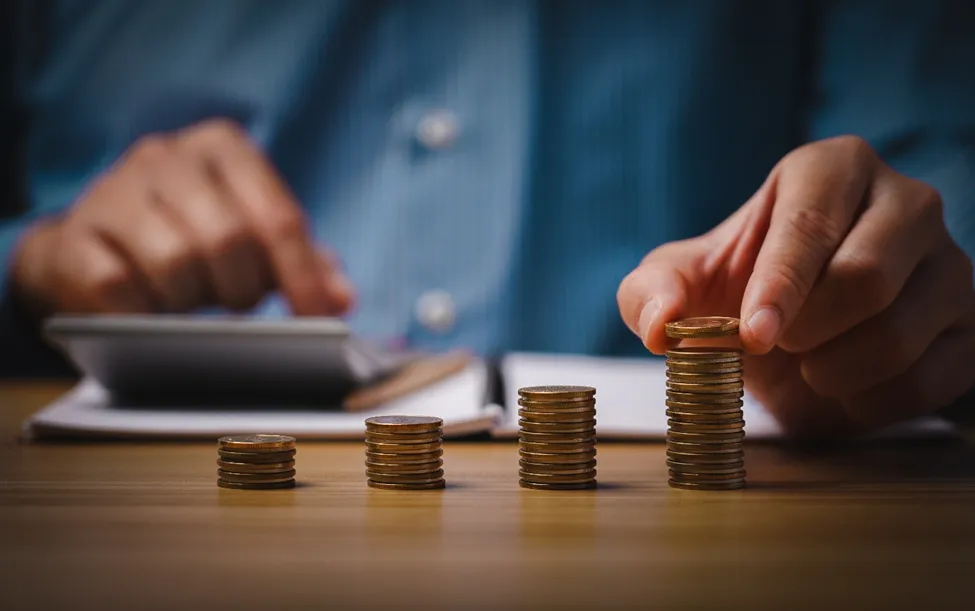 A person stacking gold coins into four increasing columns on a desk next to a calculator and notebook, symbolizing the distribution and growth of a class action settlement fund.