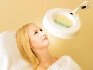 A blonde woman lying down on a white treatment bed while a bright circular magnifying lamp is positioned over her face for a skin examination.
