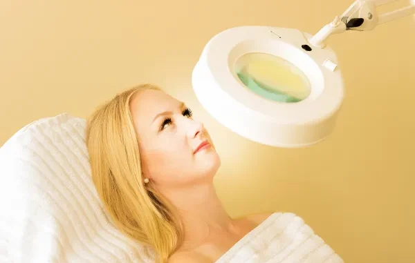 A blonde woman lying down on a white treatment bed while a bright circular magnifying lamp is positioned over her face for a skin examination.