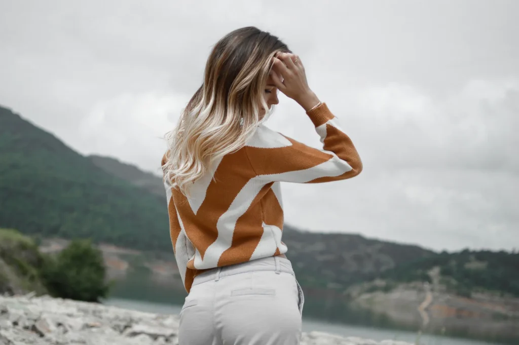 A woman from behind wearing a thick, mustard yellow and white chevron-patterned knit sweater while standing in front of a scenic mountain and lake backdrop.