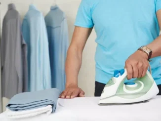 A close-up of a person in a blue t-shirt using a modern steam iron on a crisp white shirt, with freshly pressed shirts hanging in the blurred background.