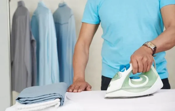A close-up of a person in a blue t-shirt using a modern steam iron on a crisp white shirt, with freshly pressed shirts hanging in the blurred background.