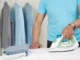 A close-up of a person in a blue t-shirt using a modern steam iron on a crisp white shirt, with freshly pressed shirts hanging in the blurred background.
