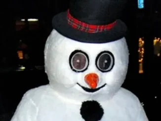 A person wearing a creepy Frosty the Snowman mascot costume with large, wide-set circular eyes, an orange carrot nose, and a black top hat with a plaid band.
