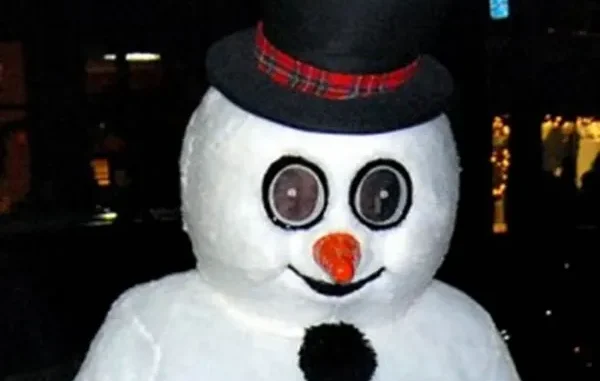A person wearing a creepy Frosty the Snowman mascot costume with large, wide-set circular eyes, an orange carrot nose, and a black top hat with a plaid band.