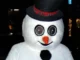 A person wearing a creepy Frosty the Snowman mascot costume with large, wide-set circular eyes, an orange carrot nose, and a black top hat with a plaid band.