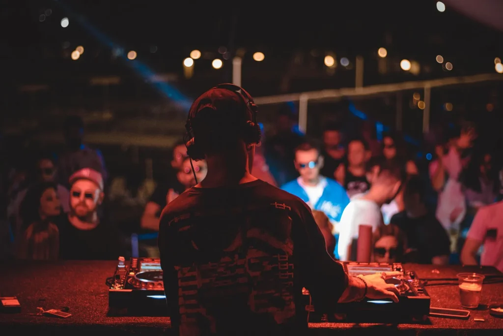 A DJ with headphones, seen from behind, performing at a crowded night venue. Warm red and orange stage lights illuminate the DJ controller and the diverse audience in the background, capturing a moment of cultural and musical exchange at a nocturnal event.