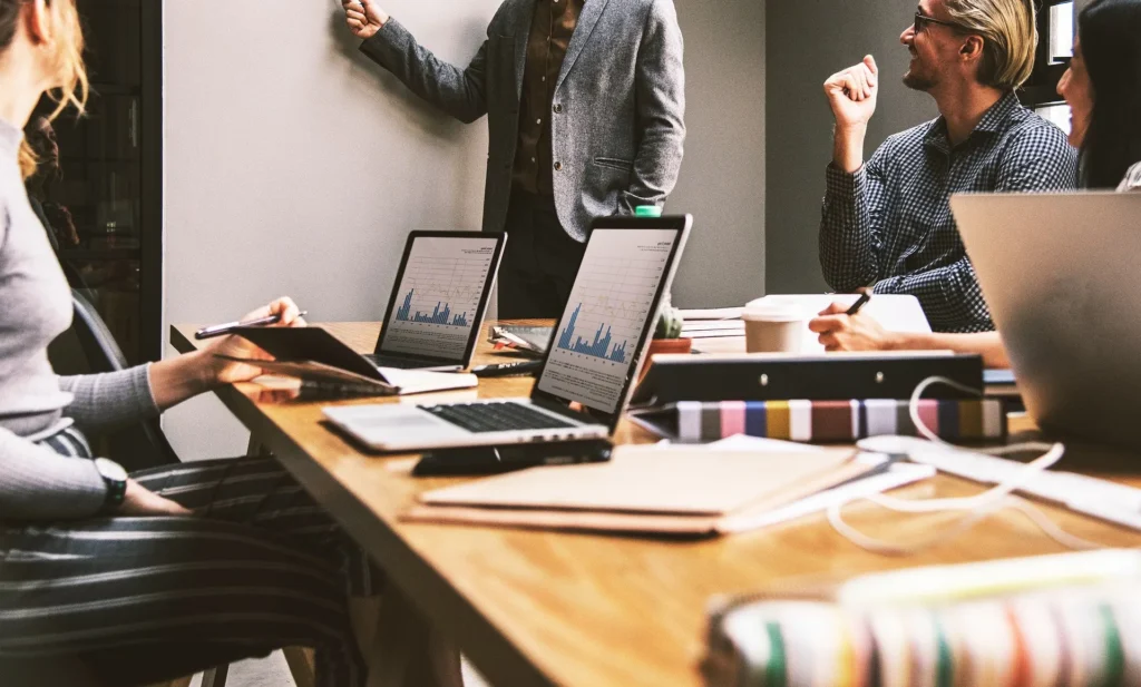 A professional group in a brightly lit office discussing data charts on laptops and a whiteboard, representing a local business team analyzing lead generation strategies.