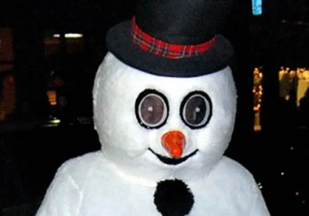 A close-up, slightly eerie photo of a person wearing a Frosty the Snowman mascot costume. The costume features a large white head with a black top hat with a plaid band, a carrot nose, and wide, circular black eyes through which the wearer's real eyes are visible.