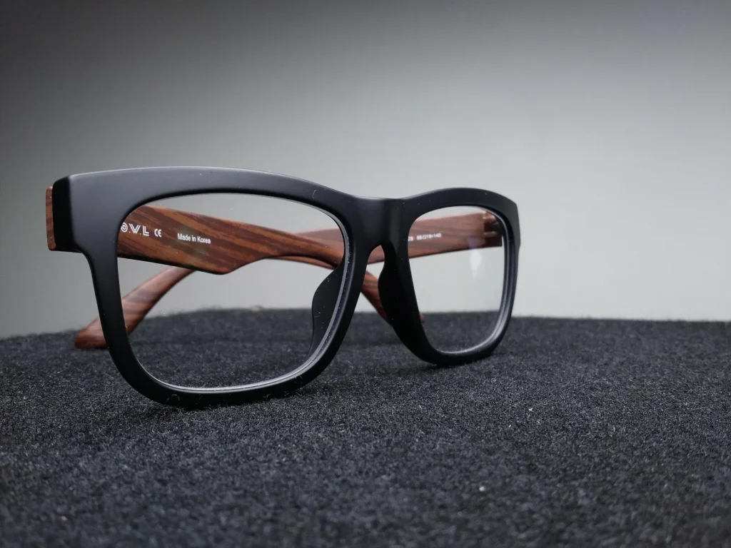 A pair of modern spectacles with matte black rectangular frames and polished wood-grain temples, resting on a dark textured surface.