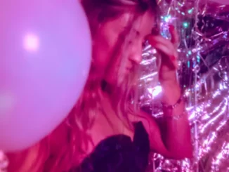 A woman in a dark patterned dress stands in a pink-lit room filled with iridescent streamers and a large white balloon, capturing the vibrant atmosphere of a nightlife event.