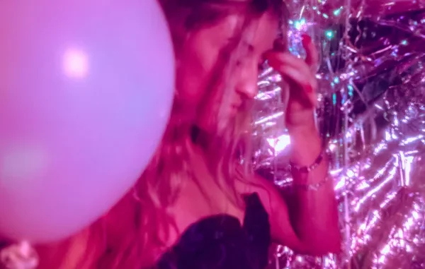 A woman in a dark patterned dress stands in a pink-lit room filled with iridescent streamers and a large white balloon, capturing the vibrant atmosphere of a nightlife event.