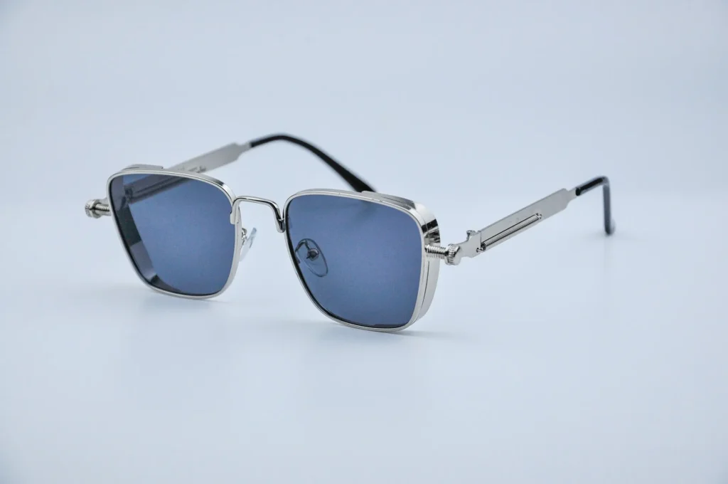 A pair of modern, square-framed sunglasses with a polished silver metal finish and dark blue-tinted lenses. The frames feature industrial-style screw details at the temples and a sleek, minimalist bridge, set against a clean, neutral background.