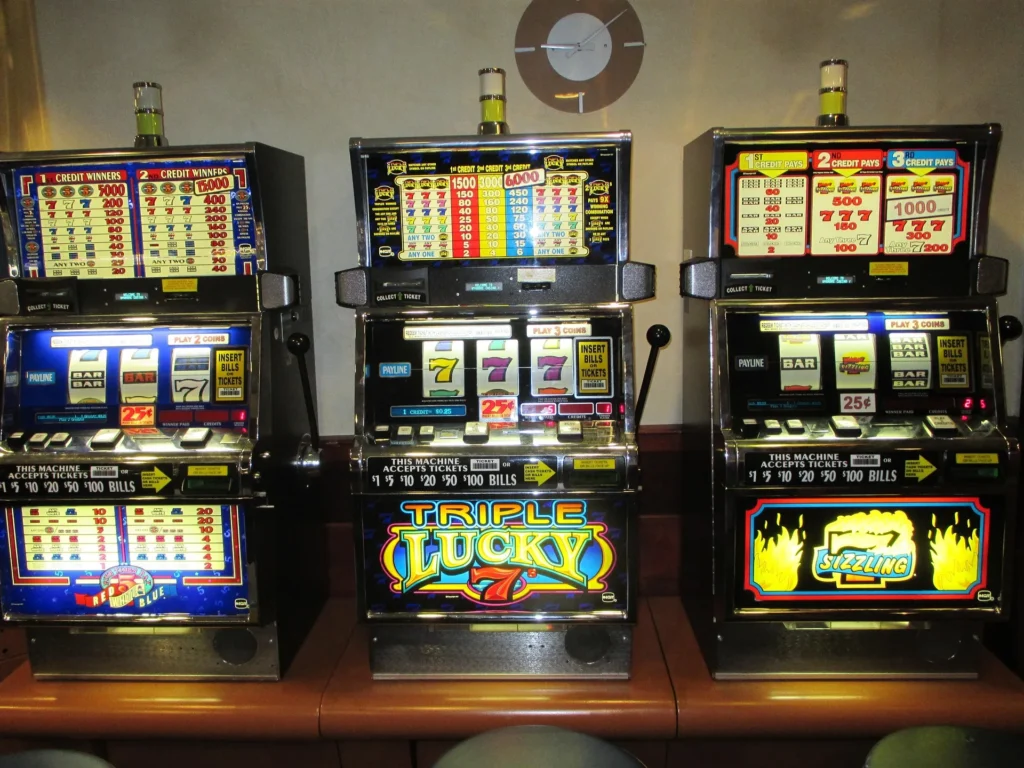 Three classic three-reel mechanical slot machines lined up on a casino floor, featuring traditional symbols like 7s and bars with physical payout tables displayed above the reels.