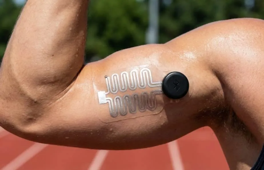 A close-up of a flexible, transparent biometric sensor patch adhered to an athlete's bicep. The patch features serpentine conductive traces and a small black circular transmitter, illustrating modern wearable EMG or ECG technology in action on a running track.