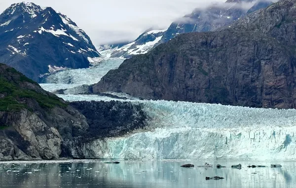 A wide view of a bright blue glacier flowing down between steep, rocky mountains into a calm bay with small floating icebergs.