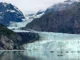 A wide view of a bright blue glacier flowing down between steep, rocky mountains into a calm bay with small floating icebergs.