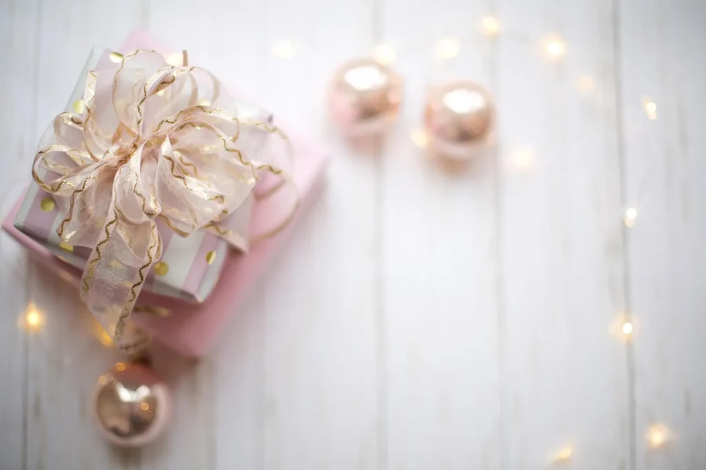 An elegant stack of pink and white gift boxes topped with a voluminous, shimmering gold-edged ribbon. The presents sit on a white wooden surface, surrounded by soft-focus rose gold ornaments and warm twinkling fairy lights, creating a cozy and romantic atmosphere.