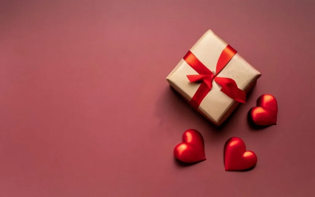 A top-down view of a small, brown paper gift box tied with a vibrant red ribbon bow, placed on a deep mauve background. Three glossy, 3D red hearts are scattered near the gift, creating a romantic and minimalist aesthetic.