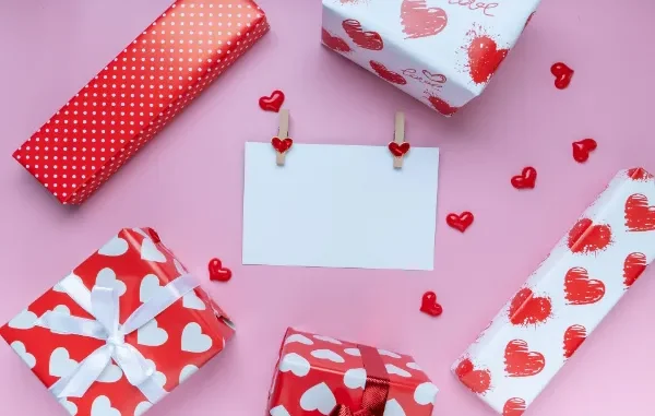Top-down view of five red and white heart-patterned gift boxes surrounding a blank white note card held by heart-shaped clothespins on a pink surface.