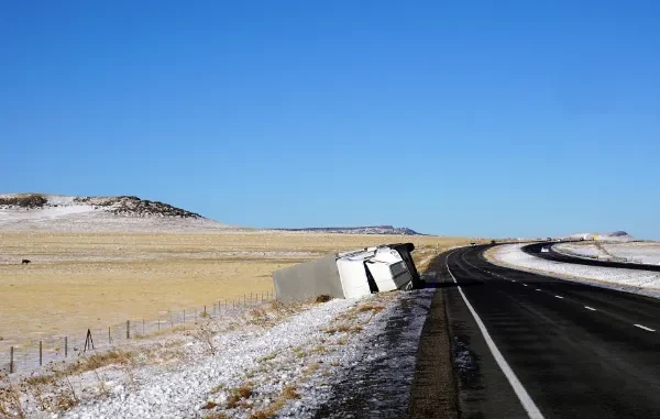 A large white delivery semi-truck overturned on its side on a snowy roadside with a rural, hilly landscape in the background.