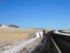 A large white delivery semi-truck overturned on its side on a snowy roadside with a rural, hilly landscape in the background.