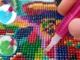 A close-up of a pink ergonomic diamond painting pen being used to place a green resin drill onto a colorful canvas. Two circular insets show the pen dipping into blue wax and picking up drills from a tray.