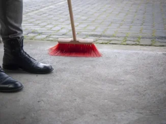 Close-up of a person in black boots using a red-bristled broom to sweep a concrete garage floor and driveway to improve curb appeal.