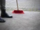 Close-up of a person in black boots using a red-bristled broom to sweep a concrete garage floor and driveway to improve curb appeal.
