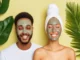 A smiling man and woman with clay face masks applied, framed by tropical green leaves against a soft yellow background, representing a holistic approach to self-care and wellness.