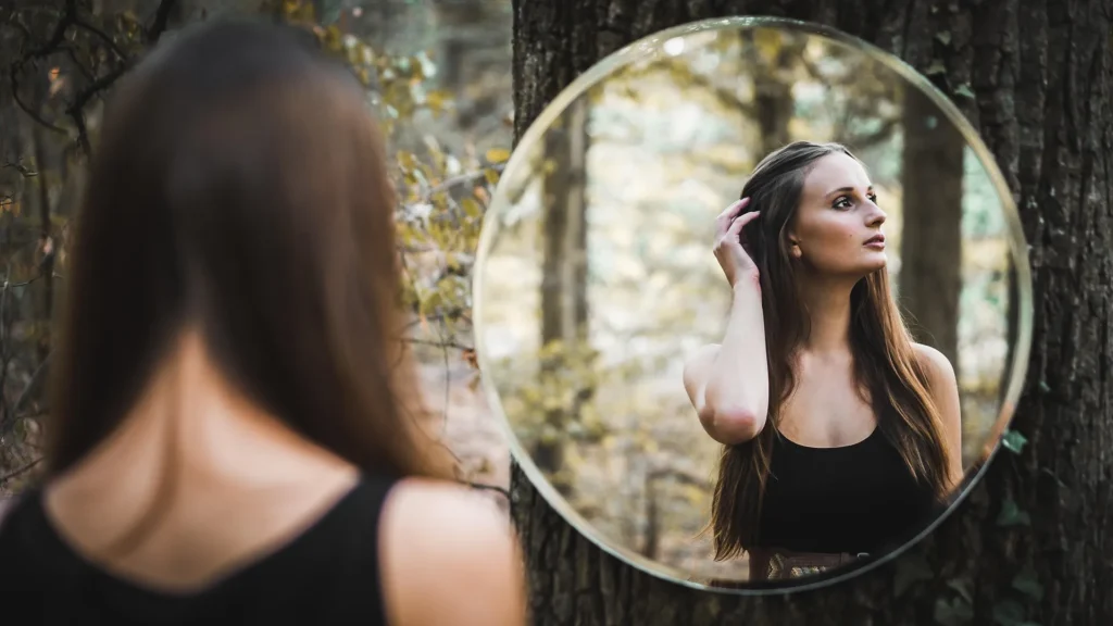 A woman stands in a forest looking into a large round mirror mounted on a tree, reflecting her profile as she tucks her hair behind her ear.