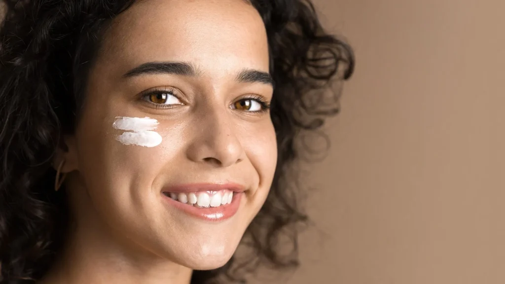 Close-up of a smiling woman with curly hair and two streaks of white facial cream on her cheek, illustrating how to feel good in your skin.