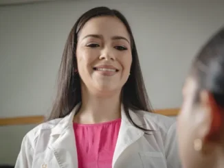 A female doctor in a white lab coat and pink blouse smiling warmly while looking down at a patient during a medical consultation.