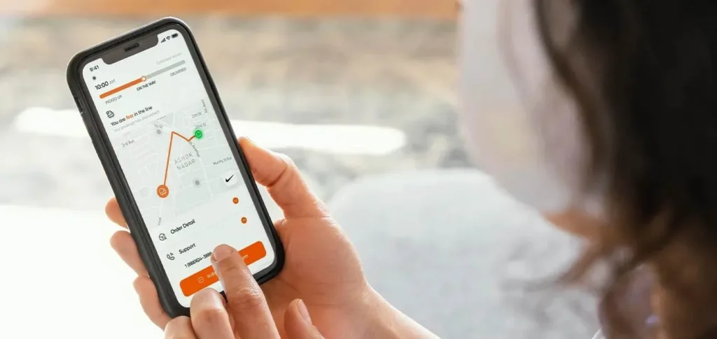 A close-up of a person holding a smartphone showing a real-time delivery tracking map. The screen displays a package "On the Way" through an urban neighborhood, highlighting the final mile delivery process.