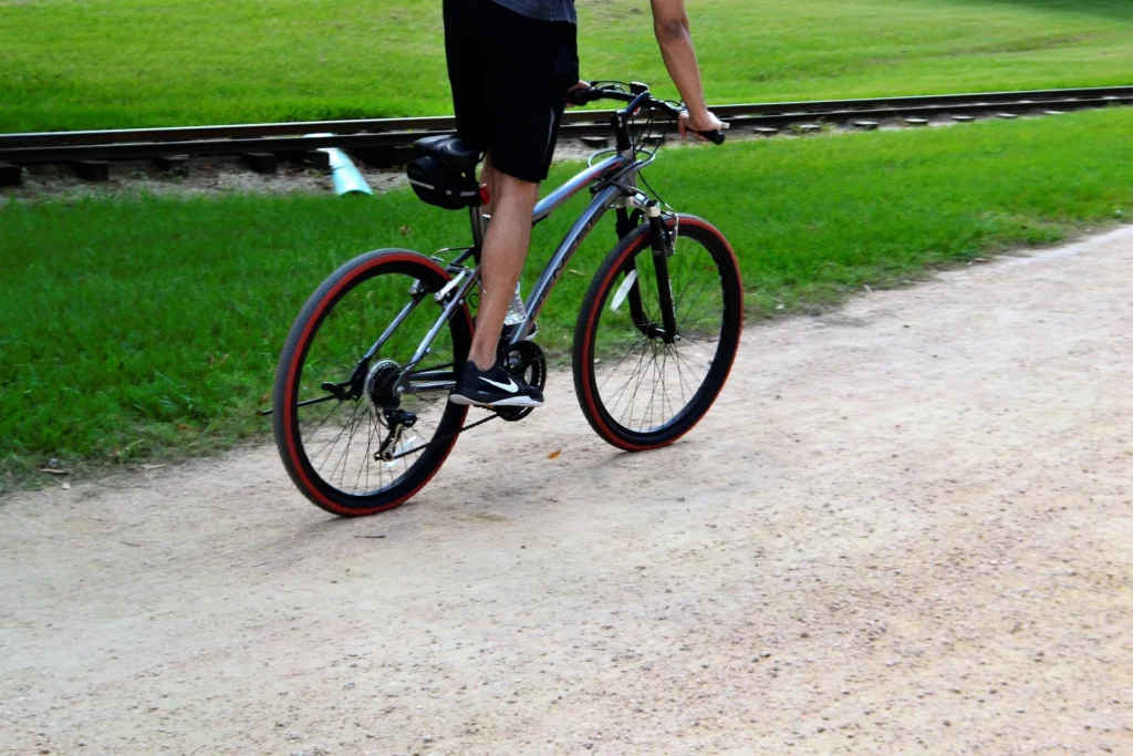 A person riding a silver mountain bike with red-rimmed tires on a gravel path next to a grassy field and train tracks, illustrating the intersection of outdoor adventure and modern lifestyle tech.