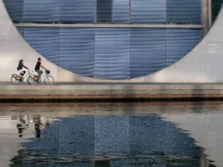 Two people riding bicycles along a waterfront path in front of a massive, modern concrete building with a large circular window and blue louvers, reflected in the water.
