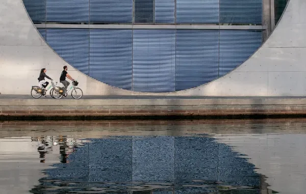 Two people riding bicycles along a waterfront path in front of a massive, modern concrete building with a large circular window and blue louvers, reflected in the water.