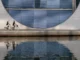 Two people riding bicycles along a waterfront path in front of a massive, modern concrete building with a large circular window and blue louvers, reflected in the water.