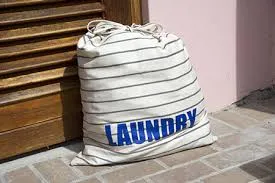 A striped canvas laundry bag labeled "LAUNDRY" sitting on a tiled floor outside a wooden door, ready for doorstep pickup.