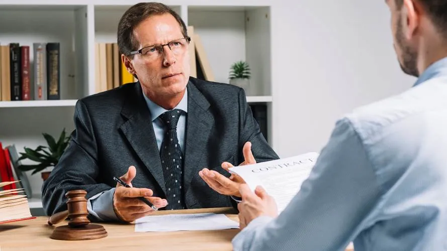 A male lawyer in a grey suit gestures toward a contract while discussing a legal case with a client across a desk.