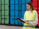 A female logistics manager in a high-visibility safety vest holding a clipboard while standing in front of green, blue, and red shipping containers.