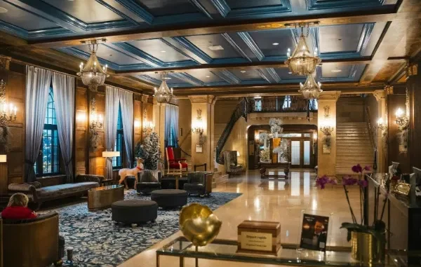 A luxurious hotel lobby featuring wood-paneled walls, blue coffered ceilings, ornate chandeliers, and a grand staircase decorated for the holidays.