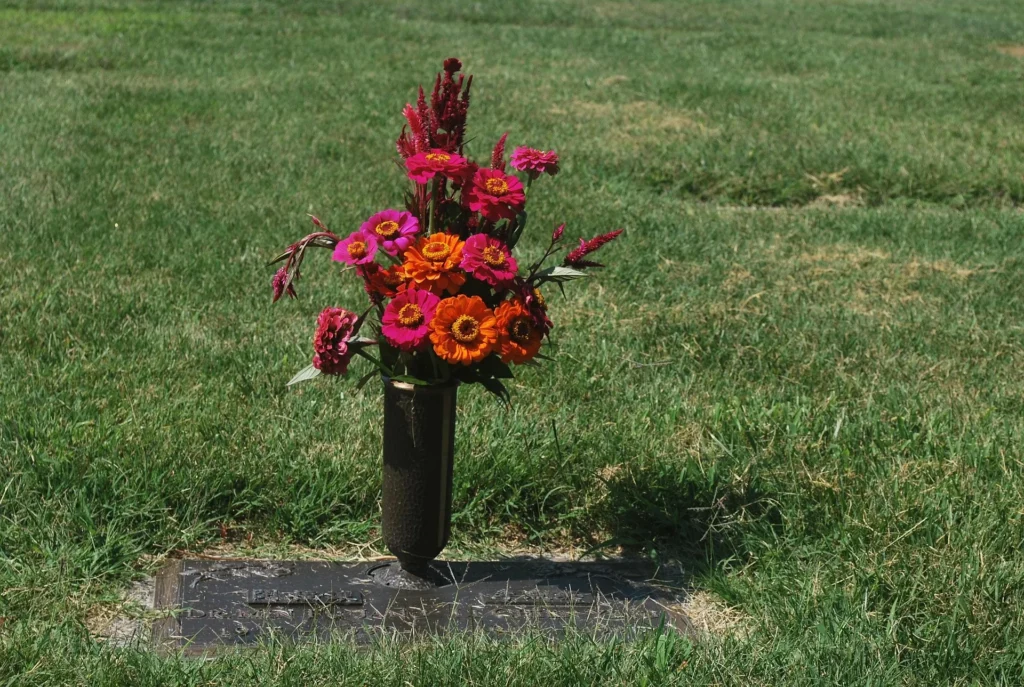 A bronze flat grave marker in a green lawn with a vase holding vibrant pink and orange zinnias.