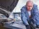 A focused mechanic in blue overalls leaning over an open car hood, inspecting the engine components and electrical systems for a power failure diagnosis.