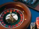 A high-angle, close-up shot of a spinning wooden roulette wheel with a white ball in motion, next to stacks of poker chips on a blue felt table.