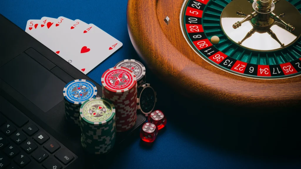 A high-angle shot featuring a laptop keyboard next to a wooden roulette wheel, stacks of poker chips, red dice, and a straight flush of playing cards on a blue surface, representing the blend of online and traditional gambling.